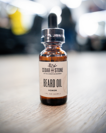 A small amber glass dropper bottle of Cedar & Stone Beard Oil with a black rubber pipette and silver cap, standing on a light wood surface with a blurred indoor background.