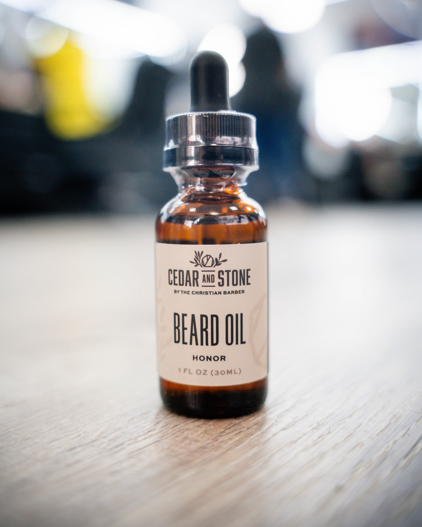 A small amber glass dropper bottle of Cedar & Stone Beard Oil with a black rubber pipette and silver cap, standing on a light wood surface with a blurred indoor background.